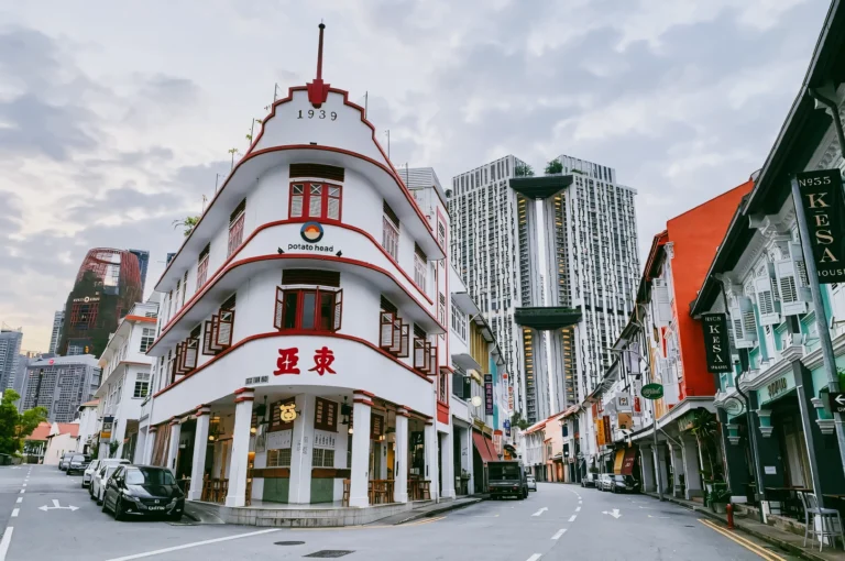 Low-angle wide shot of curved heritage shophouse on Keong Saik Road, Singapore, with red-trim facade, early morning light, and modern skyscrapers in background highlighting old vs new cityscape