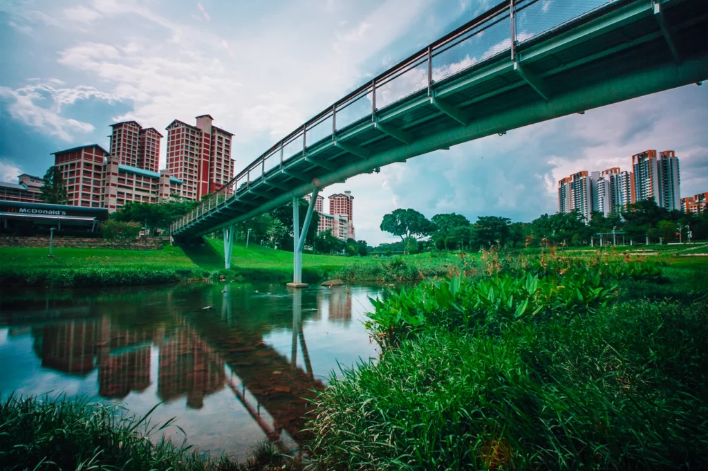 Low-angle wide shot of pedestrian bridge over canal in Singapore heartland with HDB flats and greenery, showcasing peaceful community living beyond the city centre