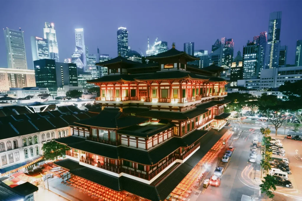 High-angle wide shot of Buddha Tooth Relic Temple in Chinatown Singapore illuminated at night with modern skyscrapers in the background, showcasing the blend of traditional Chinese architecture and modern urban skyline