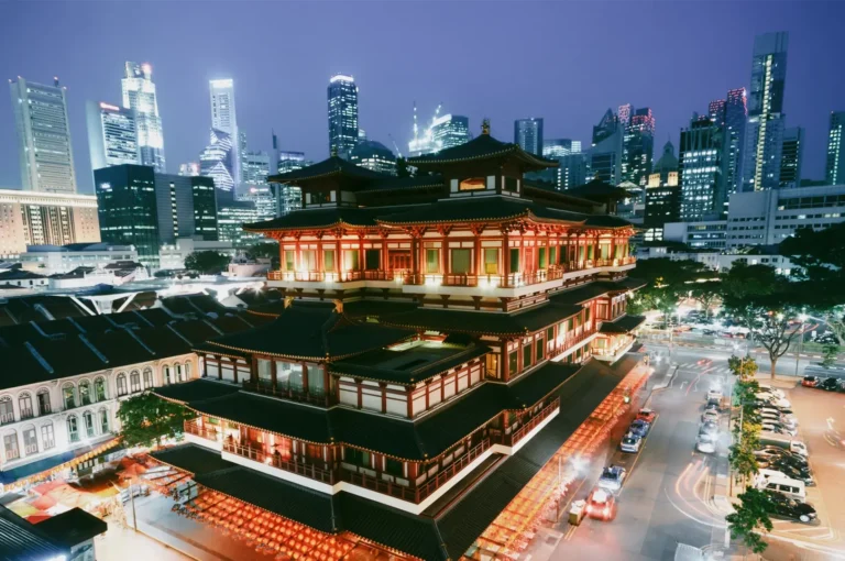 High-angle wide shot of Buddha Tooth Relic Temple in Chinatown Singapore illuminated at night with modern skyscrapers in the background, showcasing the blend of traditional Chinese architecture and modern urban skyline