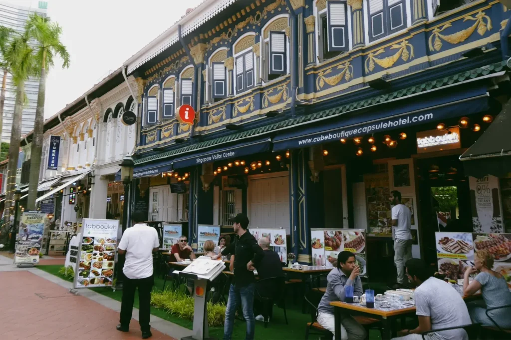 Eye-level wide street view of Kampong Glam restaurants in conserved shophouses, where diners enjoy relaxed weekend meals and communal neighborhood dining in Singapore.