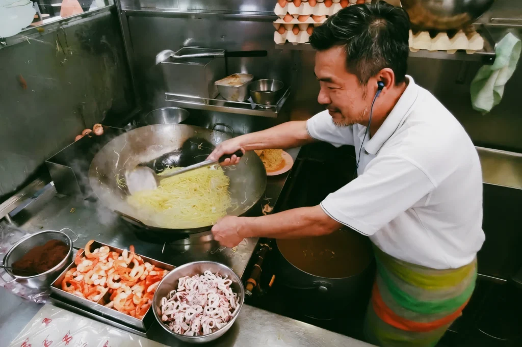 High-angle action shot of a hawker cooking yellow noodles in a large wok at a Singapore hawker stall, surrounded by fresh ingredients, steam, and metal cookware, illustrating traditional hawker food preparation.