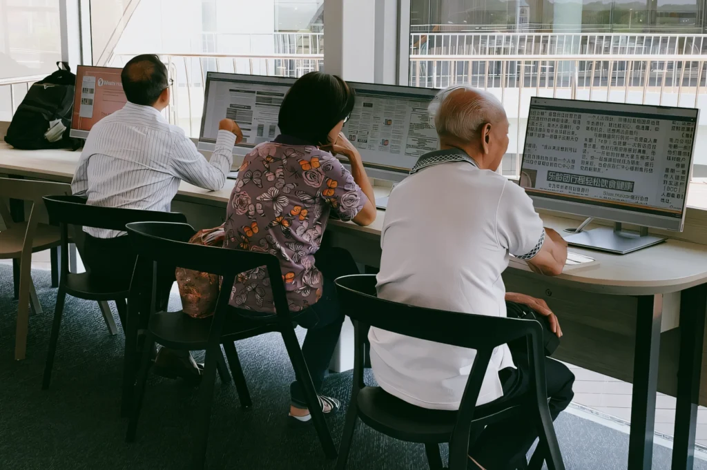 Rear eye-level medium shot of library users at desktop computers near large windows in a Singapore neighbourhood library, highlighting digital access, lifelong learning, and community engagement