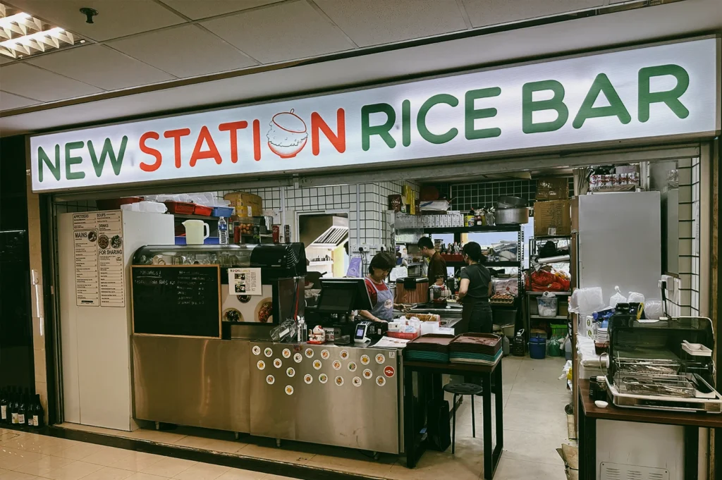 Eye-level front-facing shot of New Station Rice Bar stall at Fortune Centre, featuring hawker-style kitchen serving local Singapore dishes like salted egg chicken rice