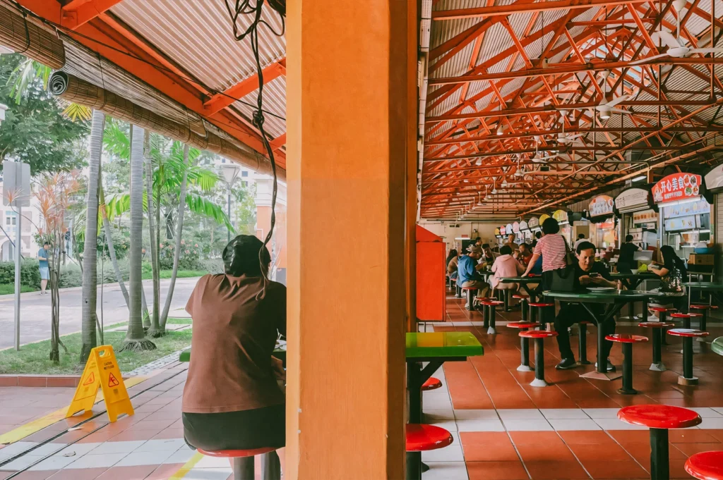 Open-air hawker centre with red stools, tiled flooring, and food stalls under a high metal roof, captured in a side perspective eye-level shot emphasizing the airy layout and everyday dining atmosphere.