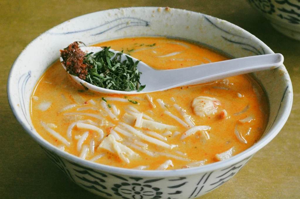 Close-up top-down shot of a bowl of Singapore laksa with coconut curry broth, rice noodles, and fresh herbs on a spoon, highlighting a classic hawker dish at a neighborhood food stall.