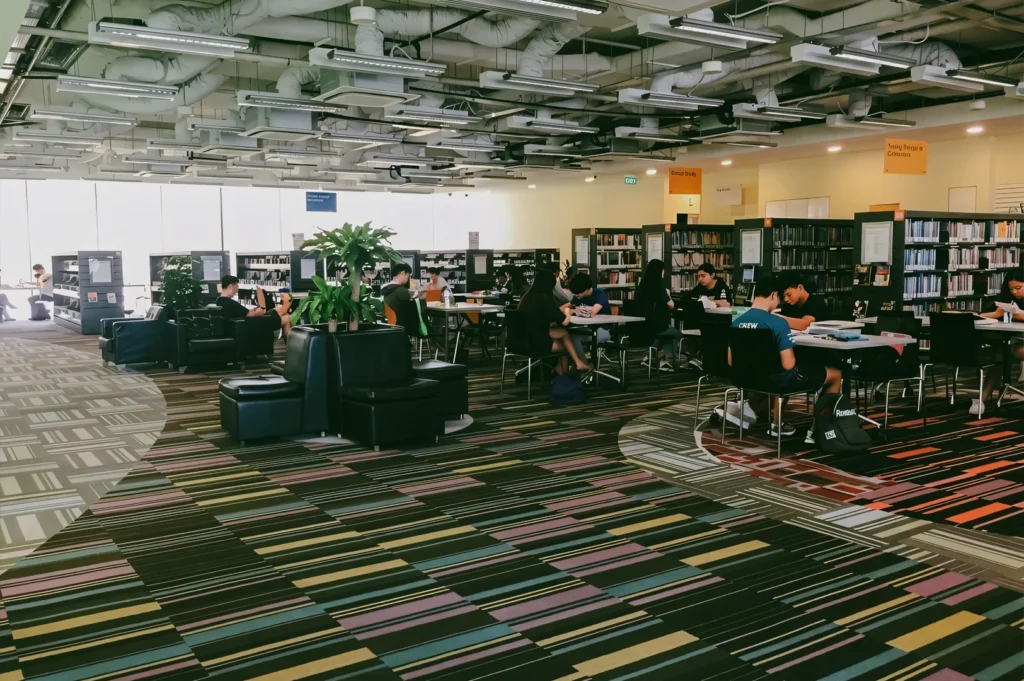 Wide-angle interior shot of busy Singapore public library reading area with study tables, bookshelves, and diverse visitors, emphasizing community learning hub and shared neighbourhood space