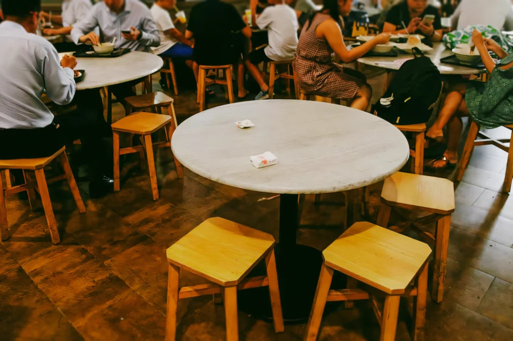 Round marble-topped hawker table with small stools and tissue packets used for seat reservation, captured in a slightly elevated angle shot showcasing traditional seating arrangements in a bustling food court.