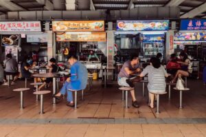 Wide‑angle eye‑level shot continuing the hawker centre scene with diners seated at tables and colorful stall signboards displaying local dishes.