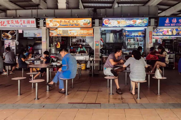 Wide‑angle eye‑level shot continuing the hawker centre scene with diners seated at tables and colorful stall signboards displaying local dishes.