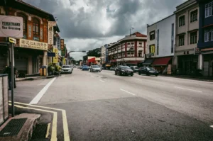 Wide-angle street-level shot of Geylang Road in Singapore, showing conserved shophouses, passing traffic, and bilingual shop signs under an overcast sky, capturing everyday urban life in the historic Geylang neighbourhood.