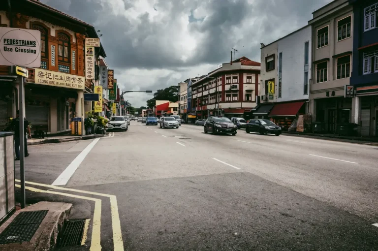 Wide-angle street-level shot of Geylang Road in Singapore, showing conserved shophouses, passing traffic, and bilingual shop signs under an overcast sky, capturing everyday urban life in the historic Geylang neighbourhood.