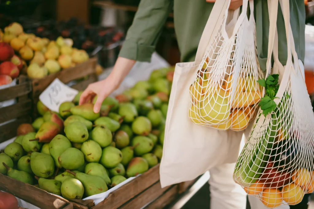 Side-angle mid-shot of a shopper using reusable mesh bags to pick fresh pears at a farmers market, emphasizing eco-friendly shopping, sustainable lifestyle, and local produce selection