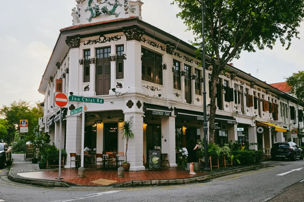 Wide-angle corner perspective of a modern café inside a restored colonial Peranakan shophouse along Joo Chiat Road, featuring ornate plaster details, timber shutters, and streetside dining in Katong Singapore.