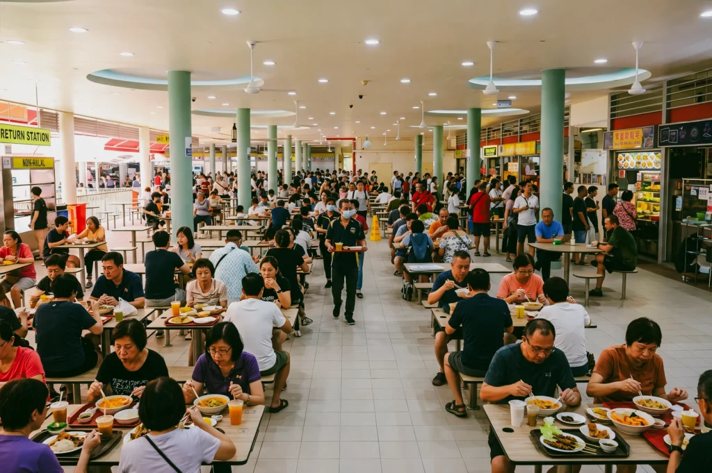 Busy Singapore hawker centre filled with diners, food stalls, and long communal tables, captured in a wide-angle eye-level shot highlighting the vibrant lunchtime crowd and local dining scene.