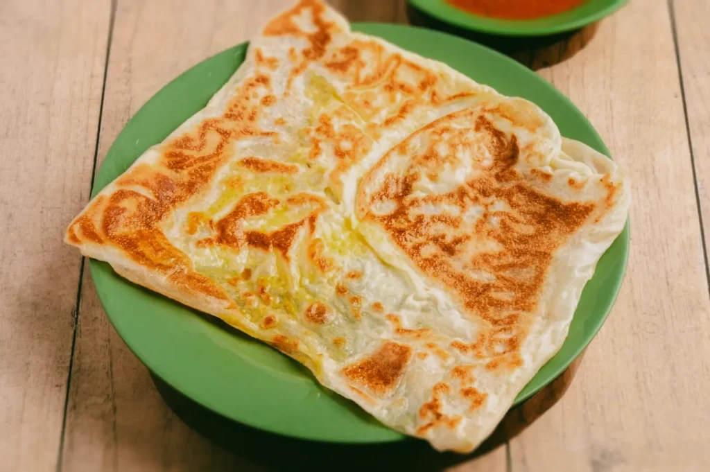 Egg roti prata on green plate in Little India Singapore, golden flaky layers with soft center, close-up overhead shot of traditional breakfast food