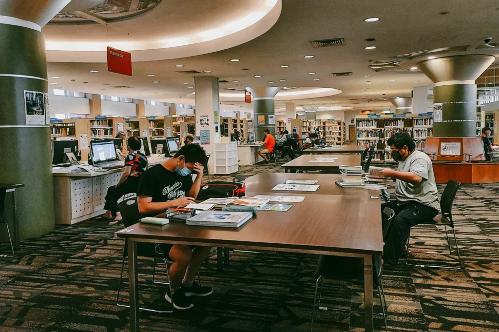Eye-level wide shot of readers studying at large tables inside a Singapore community library, illustrating quiet study environment, inclusive public space, and everyday learning activities