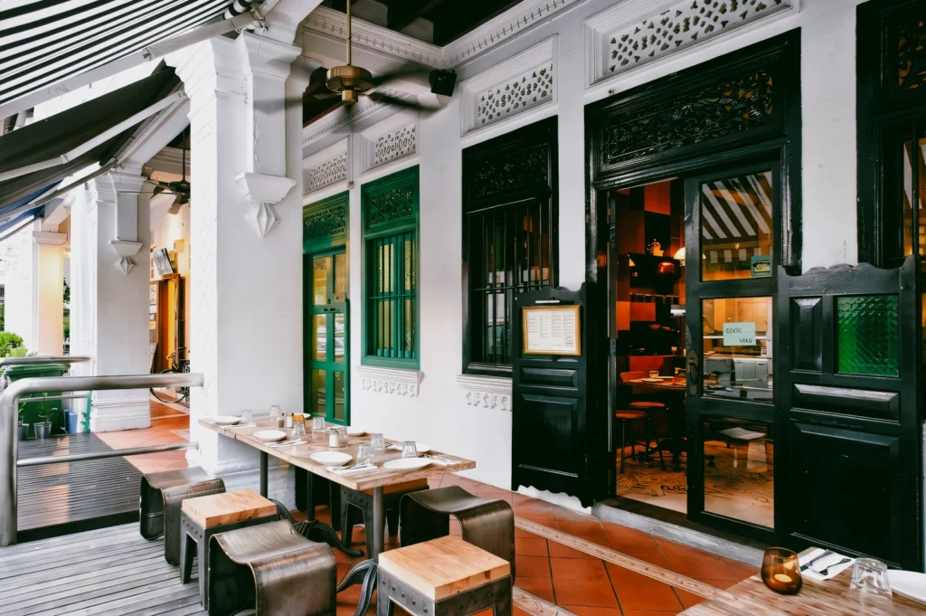 Eye-level perspective of outdoor seating at a heritage shophouse restaurant, featuring five-foot way corridor, wooden tables, and colonial-era architectural details in Singapore
