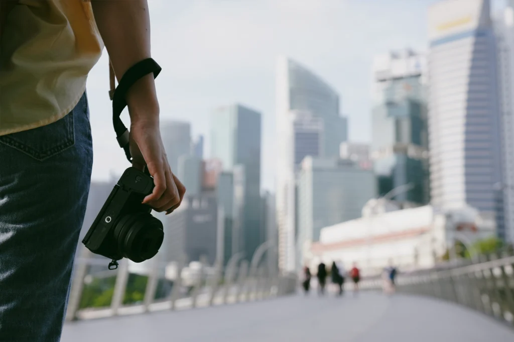 Shallow depth-of-field medium shot of a photographer holding a camera on a Singapore city walkway with urban skyline in background