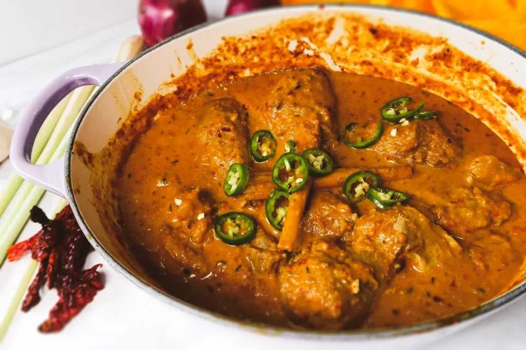 High-angle close-up of a coconut-based chicken curry simmering in a white enameled pot, featuring tender chicken in a creamy spiced sauce topped with fresh green chili slices.