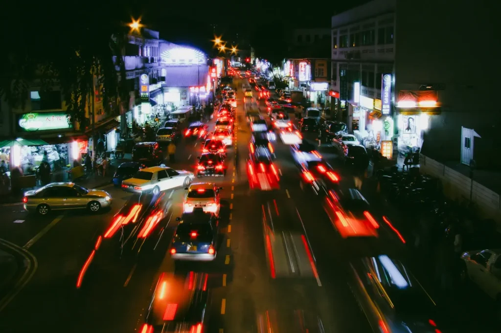 High-angle long-exposure night shot of Geylang Road in Singapore, showing busy traffic with streaking headlights, illuminated shops, and dense urban activity that reflects the nonstop energy of the Geylang district.