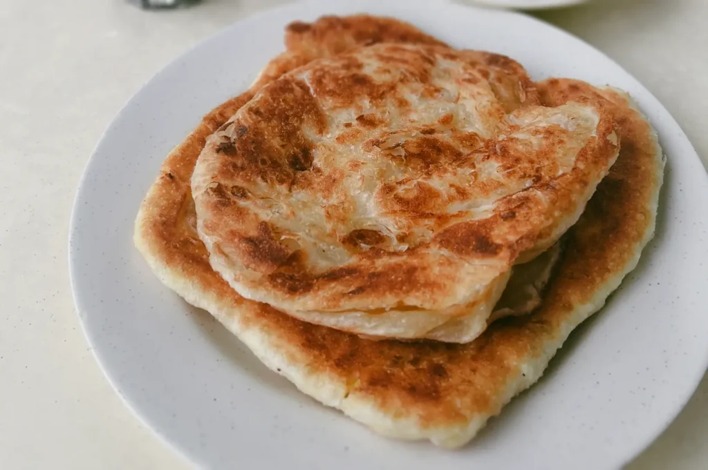 Stacked crispy roti prata Singapore on white plate showing flaky texture and golden crust, close-up eye-level food photography at prata stall