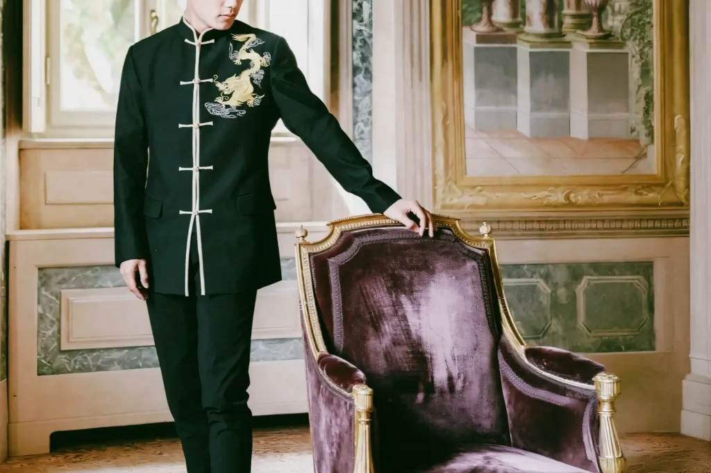 Mid-level side-angle shot of a person wearing a modern black changshan with Chinese dragon embroidery beside a classic armchair in an elegant interior, reflecting contemporary interpretations of traditional Chinese fashion in Singapore