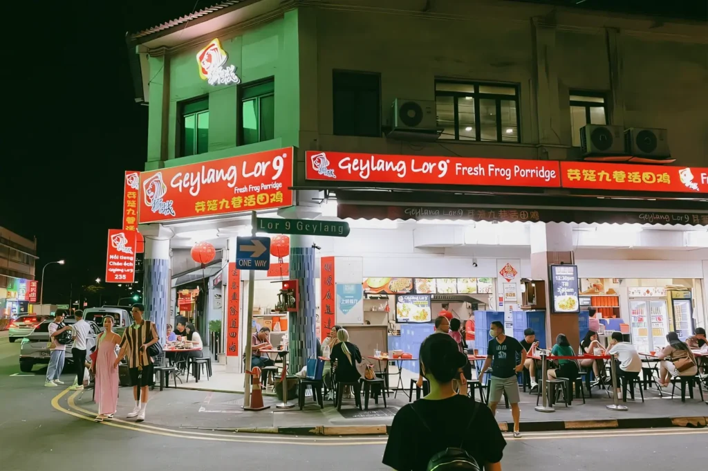 Street-level nighttime wide-angle shot of a busy Geylang lorong in Singapore, with illuminated frog porridge and late-night eateries, diners seated outdoors, and neon signage capturing Geylang’s vibrant food culture after dark.