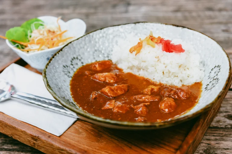 Japanese chicken curry with steamed white rice and pickled ginger served in a ceramic bowl on a wooden tray, accompanied by a side salad, captured in a slightly elevated three-quarter angle shot highlighting rich curry texture and plating presentation.