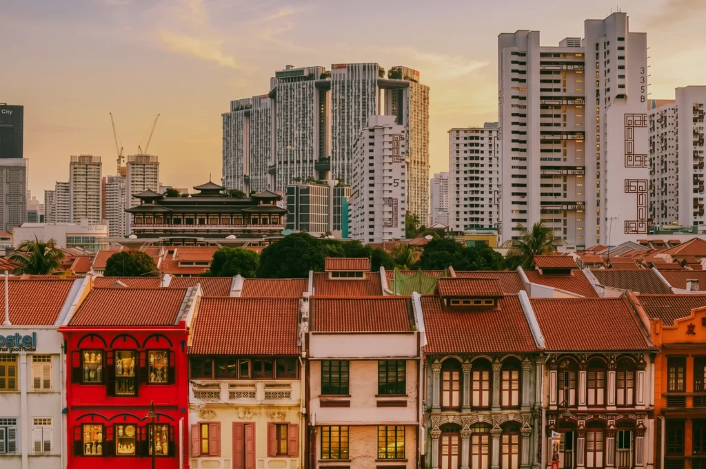 An eye-level, wide architectural shot contrasting Singapore's heritage and modernity, featuring a row of historic, brightly painted tiled-roof shophouses in Chinatown in the foreground, with the massive Pinnacle at Duxton public housing skyscrapers rising into the hazy sunset sky behind them.