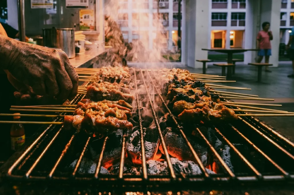 Close‑up shot of satay skewers grilling over hot charcoal, smoke rising as vendor’s hand turns skewers in lively HDB neighborhood setting.
