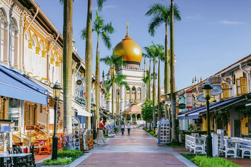 An eye-level, wide-angle shot looking down a pedestrian street in Kampong Glam lined with historic shophouses and tall palm trees, leading the eye toward the prominent golden dome of the Sultan Mosque under a clear blue sky.