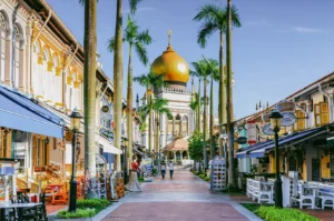 An eye-level, wide-angle shot looking down a pedestrian street in Kampong Glam lined with historic shophouses and tall palm trees, leading the eye toward the prominent golden dome of the Sultan Mosque under a clear blue sky.