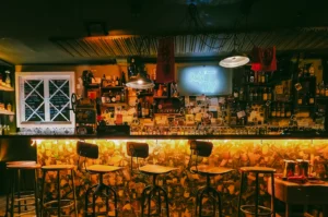 Front-facing wide shot of Sago House cocktail bar counter with high stools, backlit bottles, eclectic decor, and neon sign in a lively Duxton Hill Singapore bar