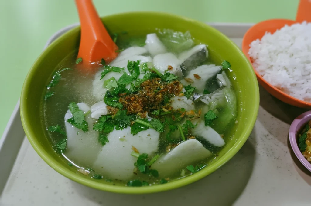 A high-angle, close-up shot of a comforting bowl of traditional sliced fish soup served in a green bowl at a Singapore hawker stall, topped with fresh coriander leaves and fried garlic, positioned on a tray next to an orange bowl of plain white rice.