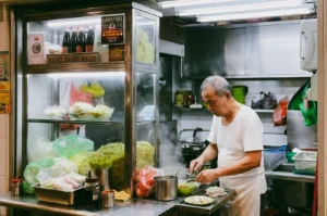 Eye-level medium shot of a traditional Singapore hawker preparing handmade fishball noodles inside a compact stainless steel stall filled with fresh ingredients at a wet market, highlighting disappearing generational recipes and authentic hawker culture