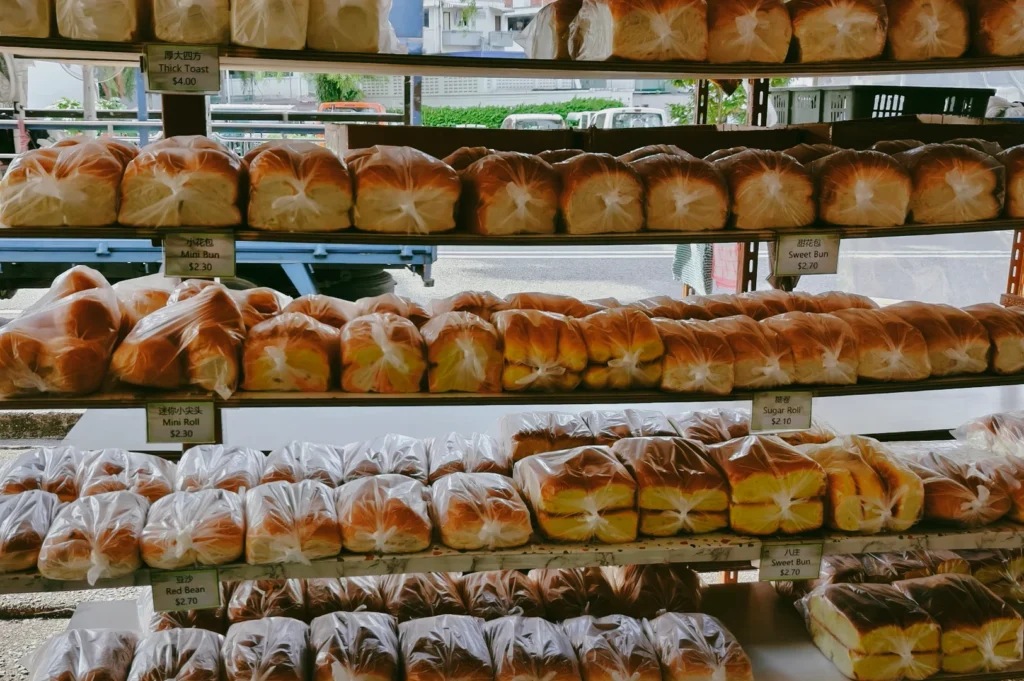 An eye-level, straight-on medium shot of a traditional Singapore neighborhood bakery's metal shelving display, filled with rows of freshly baked bread loaves, mini buns, thick toast, and sugar rolls individually wrapped in clear plastic bags with printed price tags.