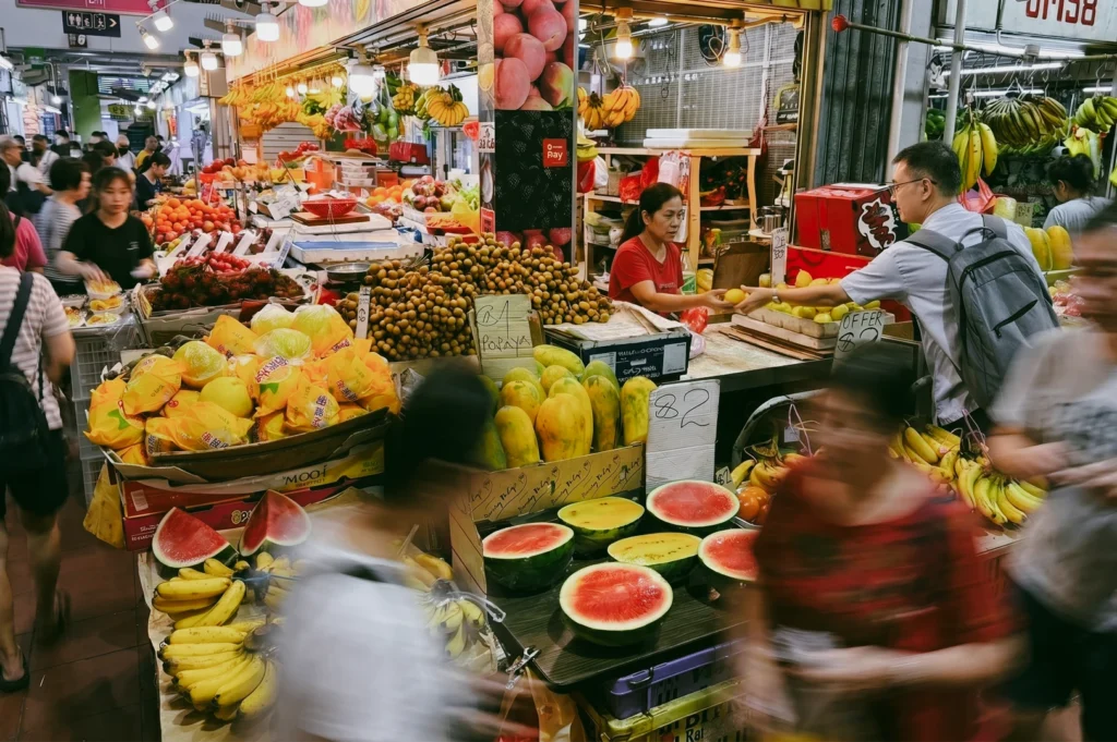 Eye-level wide-angle shot of a bustling Singapore wet market filled with colorful tropical fruits like papaya, bananas, and watermelon, as local vendors serve customers amid vibrant stalls, bright lighting, and a lively street food atmosphere showcasing authentic hawker culture.