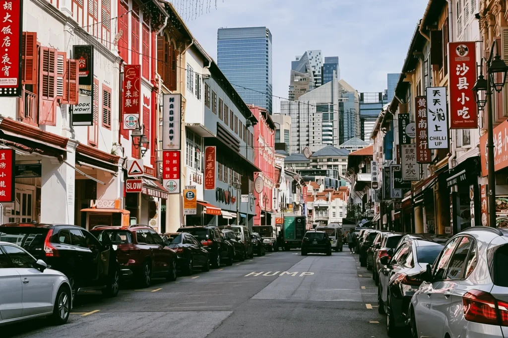 Eye-level wide-angle shot of Chinatown Singapore street with colorful heritage shophouses, parked cars, Chinese signboards, and modern skyscrapers in the background showing contrast of tradition and urban development.