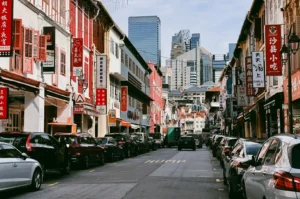 Eye-level wide-angle shot of Chinatown Singapore street with colorful heritage shophouses, parked cars, Chinese signboards, and modern skyscrapers in the background showing contrast of tradition and urban development.