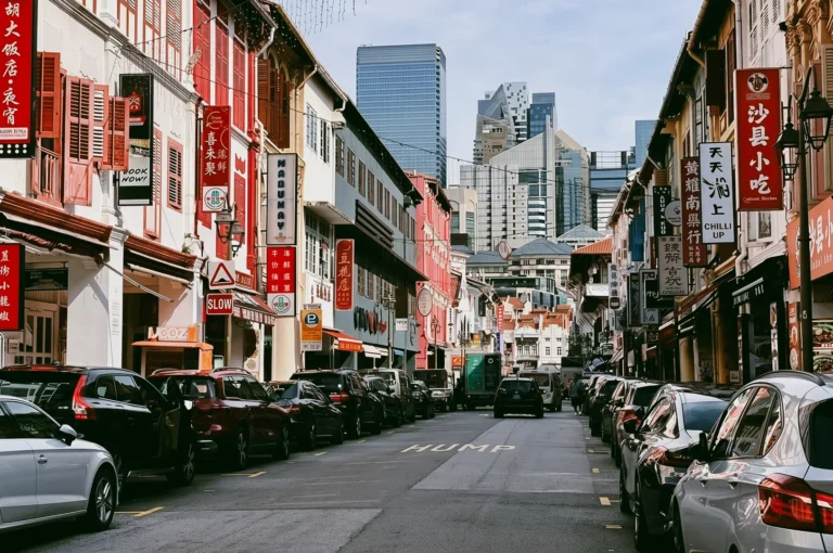 Eye-level wide-angle shot of Chinatown Singapore street with colorful heritage shophouses, parked cars, Chinese signboards, and modern skyscrapers in the background showing contrast of tradition and urban development.