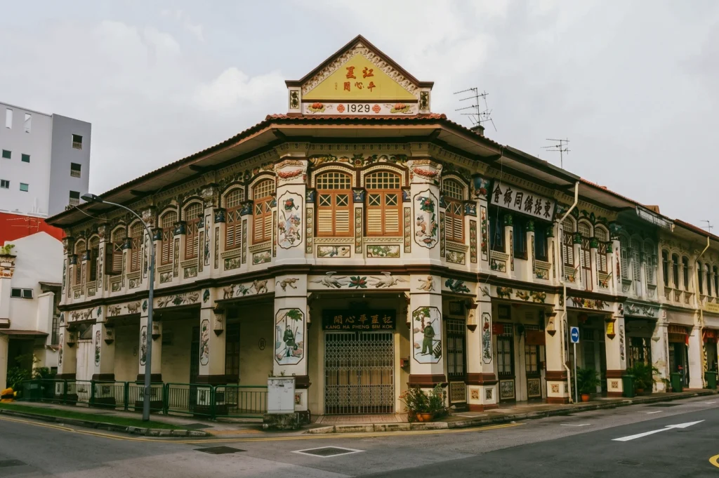 Low-angle wide shot of a restored 1929 heritage shophouse in Singapore, featuring ornate Chinese architectural details, decorative plasterwork, wooden shutters, and weathered façade at a street corner in a historic neighbourhood.
