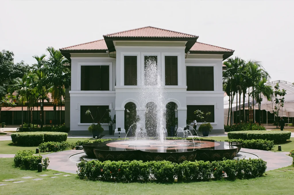 A straight-on, eye-level medium shot of the Malay Heritage Centre, a white two-story historic palace building with dark window shutters, framed by green lawns and a symmetrical water fountain spraying in the foreground.