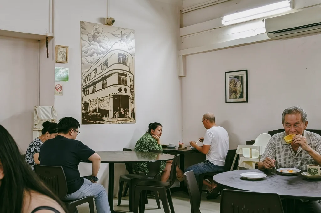Interior of traditional Singapore kopitiam with customers seated at shared tables enjoying coffee and toast – wide-angle eye-level shot capturing community dining and heritage atmosphere.