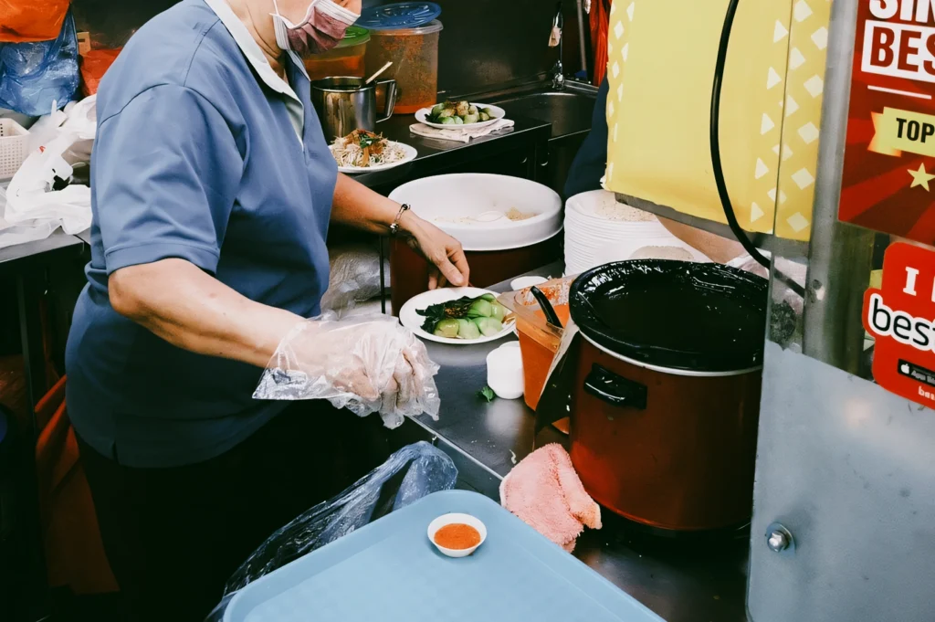 Close-up side-angle shot of a hawker preparing Hainanese chicken rice with sliced cucumbers, sauces, and rice cooker in a traditional Singapore stall, highlighting heritage recipes and everyday hawker culture