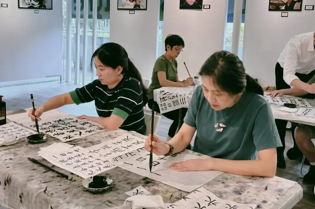 Eye-level medium shot angle of a small calligraphy class practicing Chinese brush writing on grid paper, highlighting traditional writing art, mindfulness, and community learning.