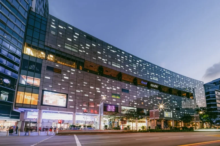 An eye-level, wide-angle exterior shot of the modern 313 Somerset mall building at dusk, featuring a dark metal facade with numerous glowing rectangular windows and bright entrance lights overlooking a street
