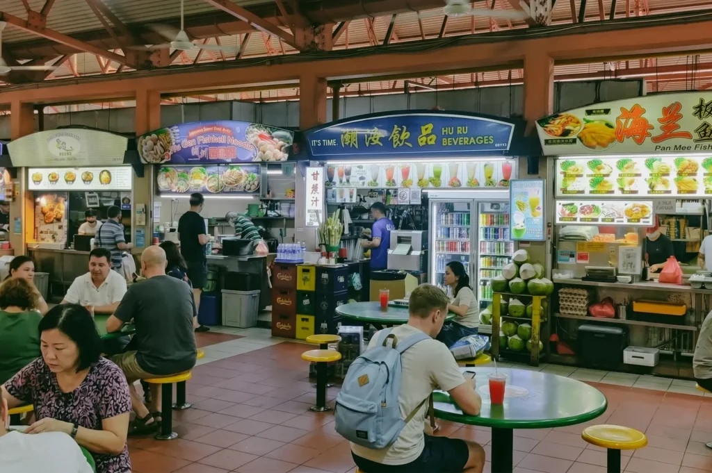 An eye-level, wide-angle shot of a vibrant row of food stalls inside a Singapore hawker centre, displaying overhead signage for fishball noodles and beverages, while a male diner wearing a blue backpack sits at a green circular table in the foreground.