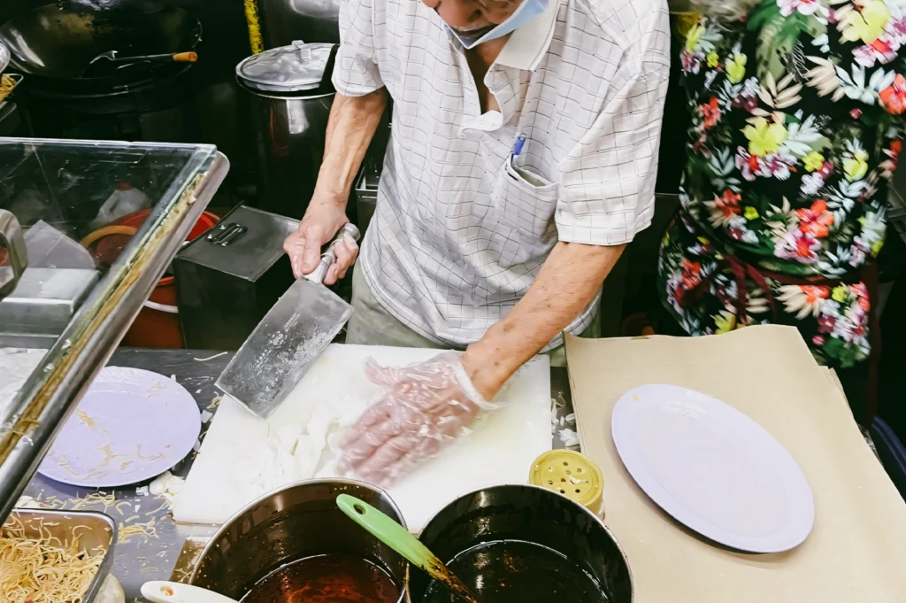 Top-down close-up shot of a hawker chopping ingredients on a cutting board with a cleaver in a busy Singapore street food stall, emphasizing manual techniques in traditional hawker cooking and fading culinary heritage