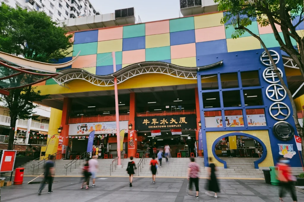 Eye-level wide shot of Chinatown Complex Singapore with colorful geometric facade, people walking into the hawker center, and signage highlighting local food culture and community life.